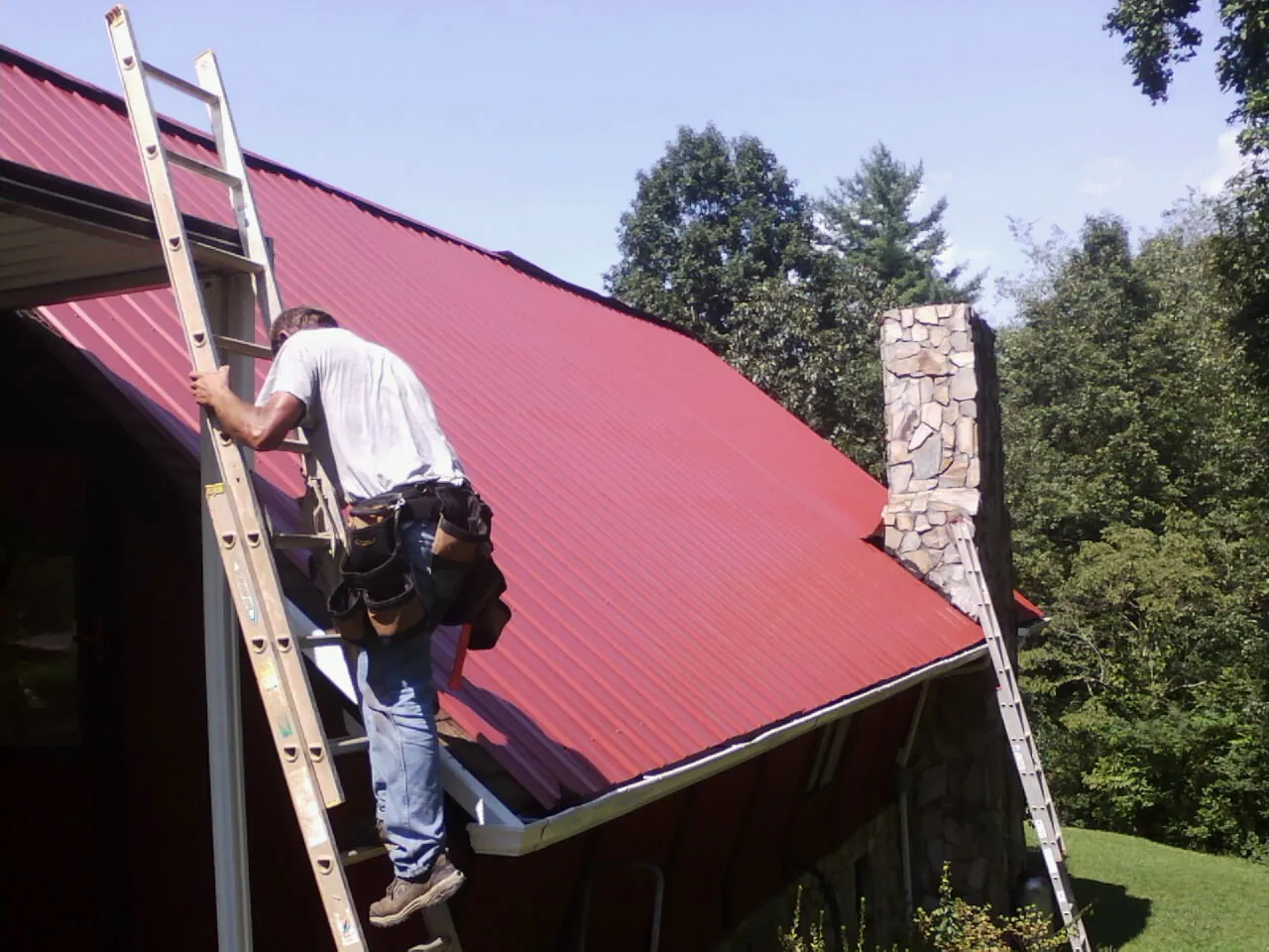 Roofer on a ladder beside a red metal roof and stone chimney — Soffit & Fascia Repair by Roofing Brothers in Belvedere Tiburon